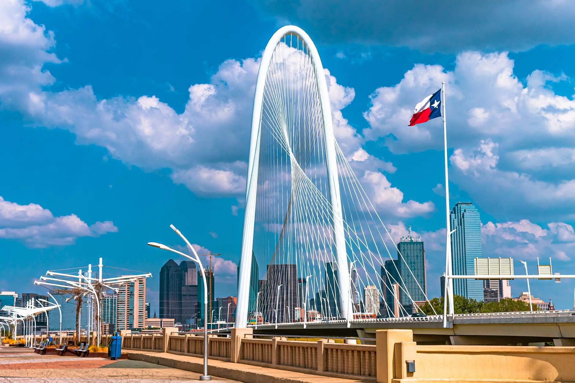 The Margaret Hunt Hill Bridge in Dallas, Texas with the Texas flag flying against a vivid blue sky and downtown skyline behind