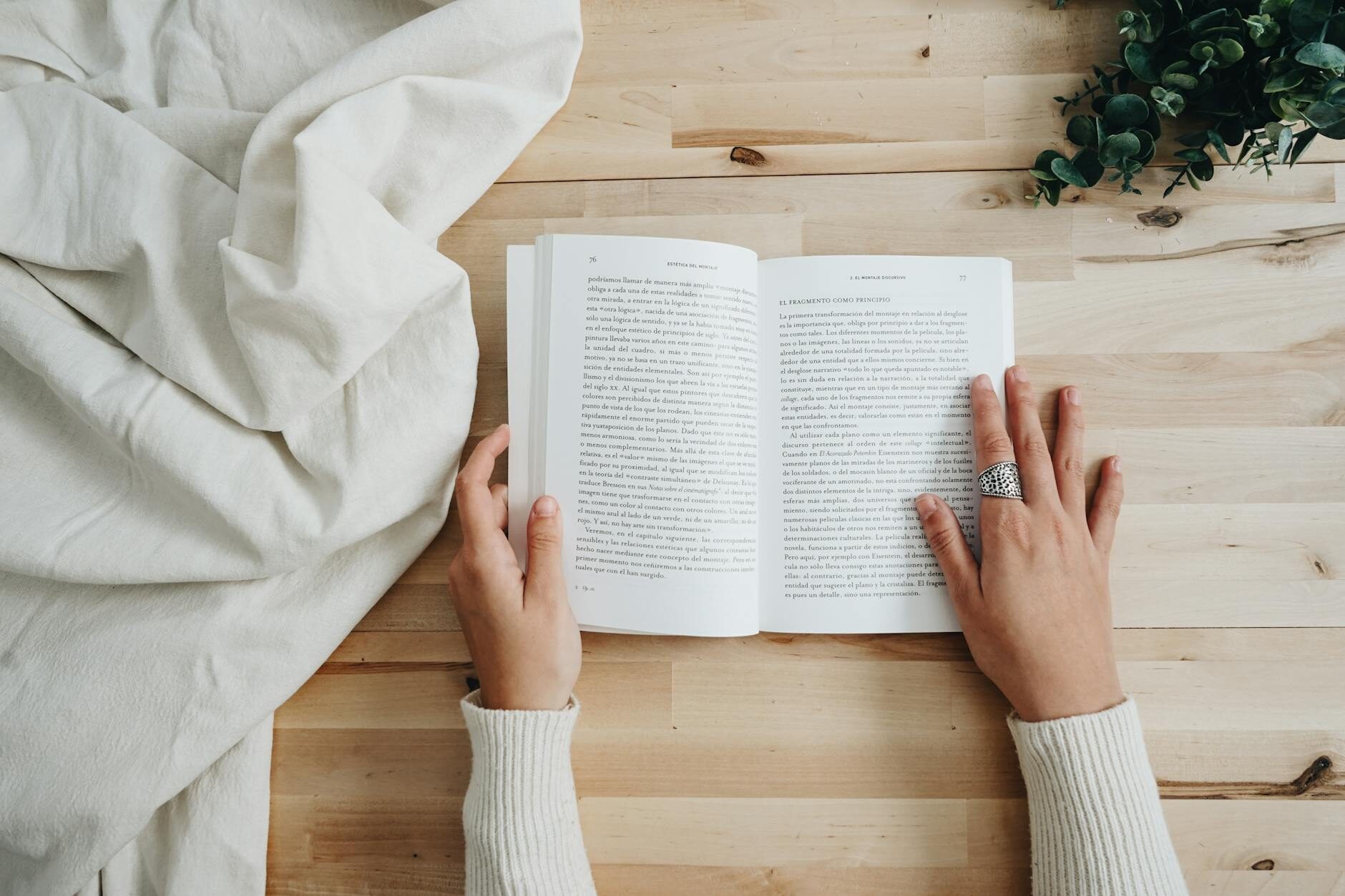 Hands holding an open book on a wooden surface with white linen and a sprig of eucalyptus