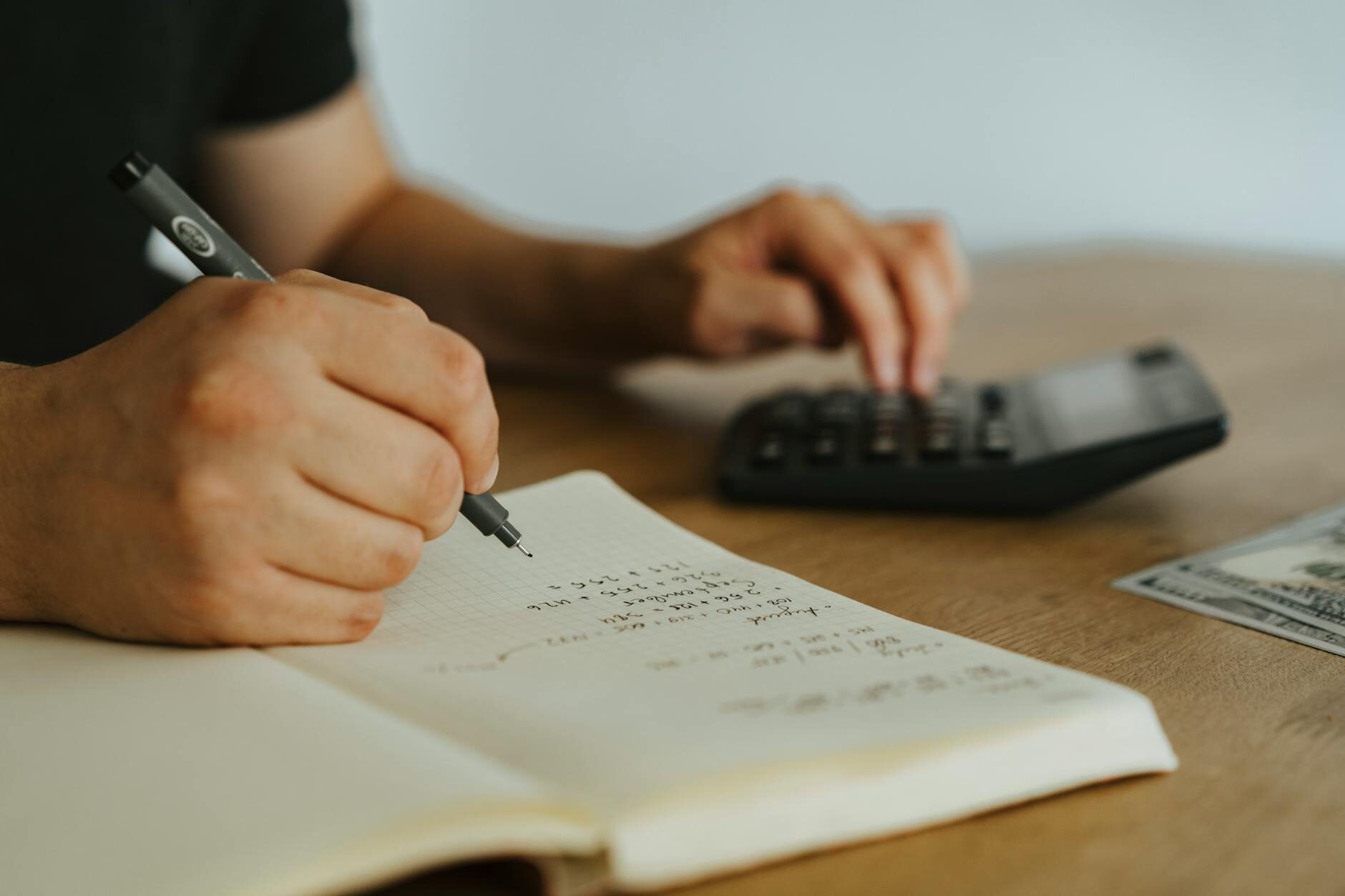 Close-up of hands writing financial calculations in a notebook with a calculator and dollar bills on a wooden desk