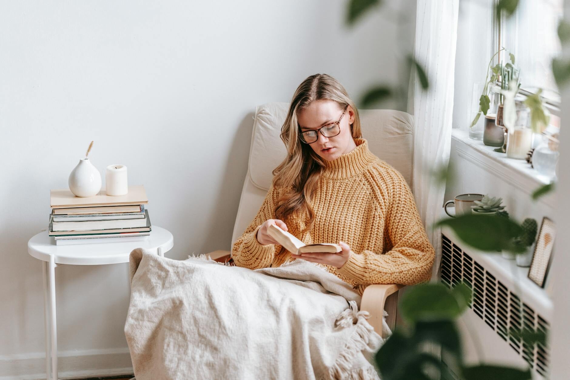 A woman in a cozy sweater reading a book in an armchair by a sunny window with plants and stacked books nearby