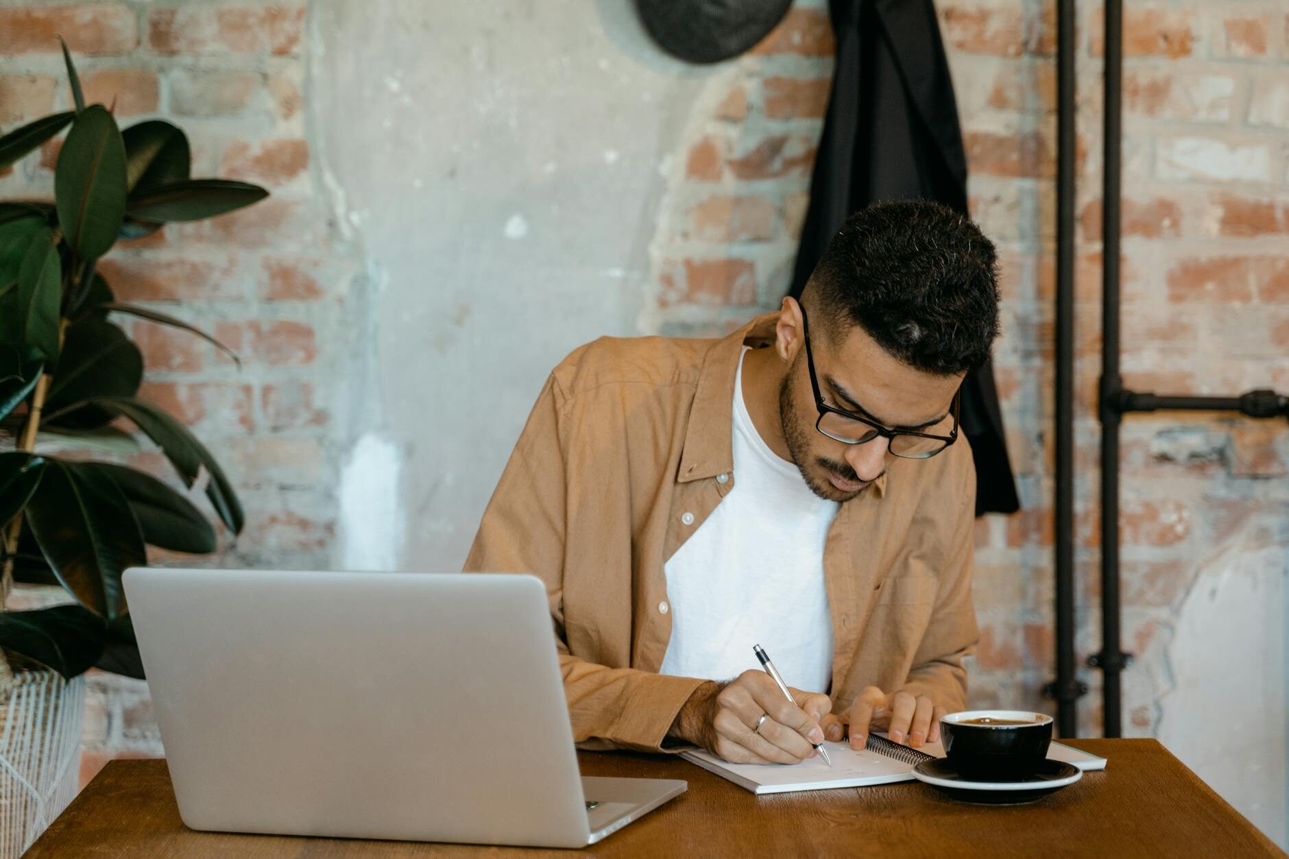 A man in a brown jacket writing in a notebook at a cafe table with coffee, brick wall behind