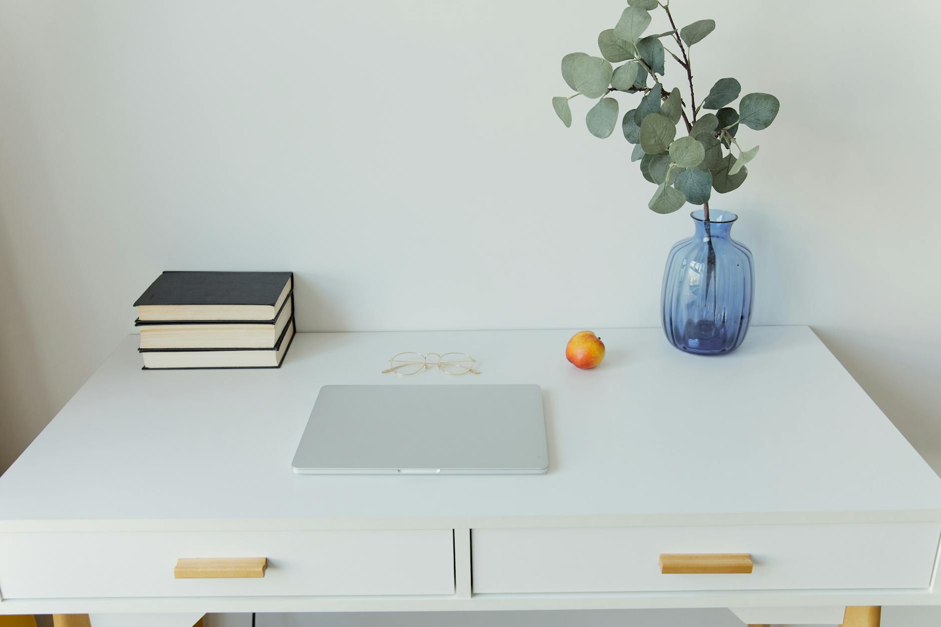 A minimalist white desk with stacked books, a silver laptop, and a glass vase with eucalyptus