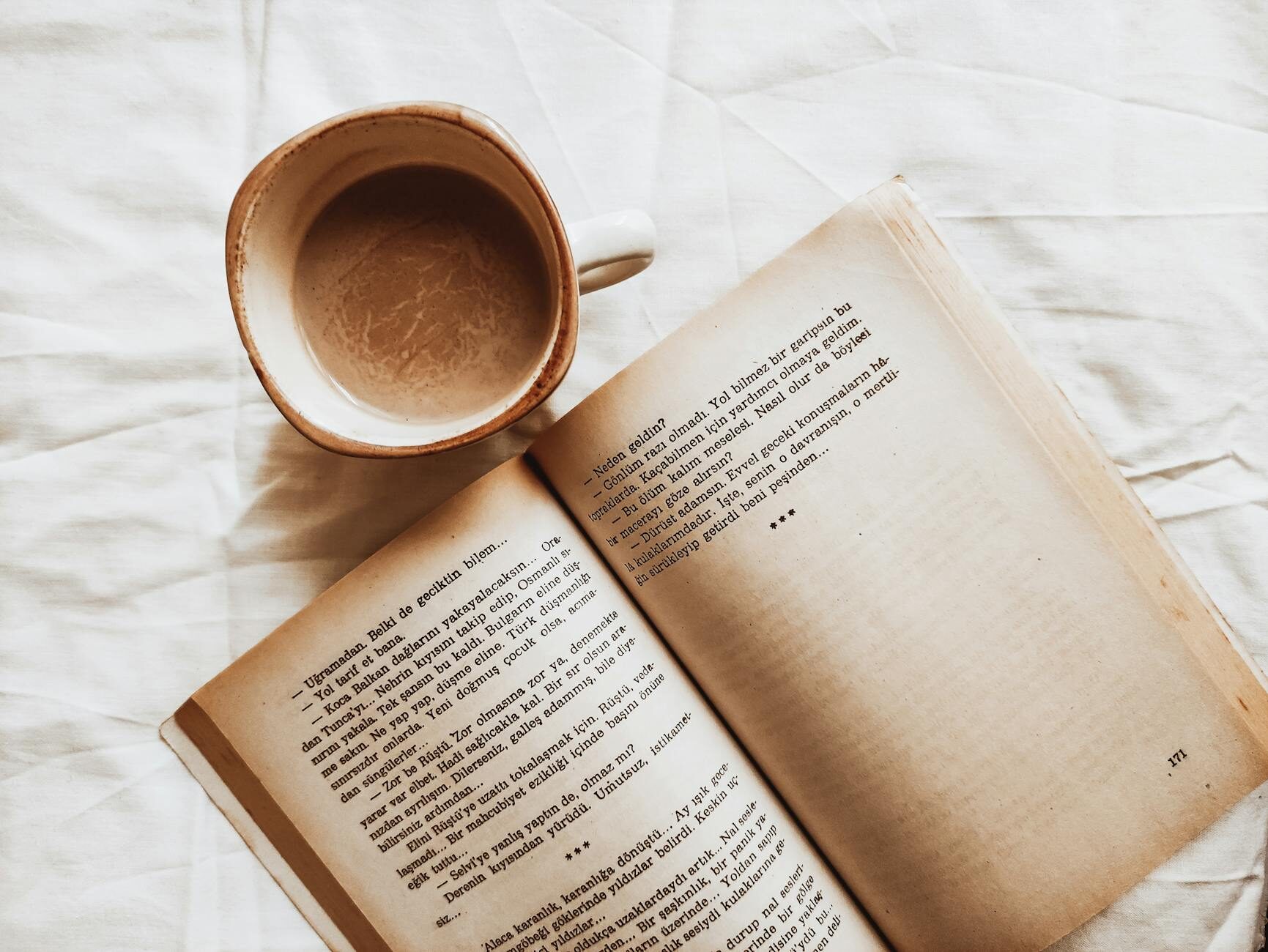 An open book beside a ceramic coffee cup on white linen in warm light